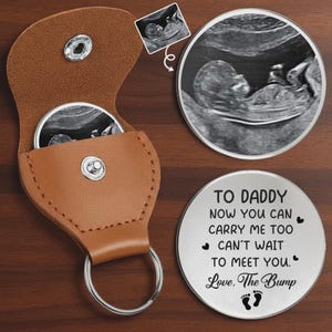 Op de afbeelding: Een bruin leren sleutelhanger met een zilveren ring, met een ronde metalen munt met een echografiebeeld. Een aparte ronde metalen munt heeft de tekst "TO DADDY NOW YOU CAN CARRY ME TOO CAN'T WAIT TO MEET YOU. Love, The Bump."
