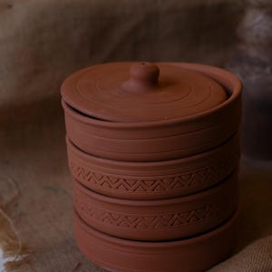 Op de afbeelding: Een stapel van vier terracotta-kleurige keramische kommen met deksels. De kommen hebben een eenvoudig, rustiek ontwerp met een getextureerd oppervlak en een decoratieve band met driehoekige patronen. Het deksel heeft een kleine knop. De kommen zijn gestapeld op een jute oppervlak.