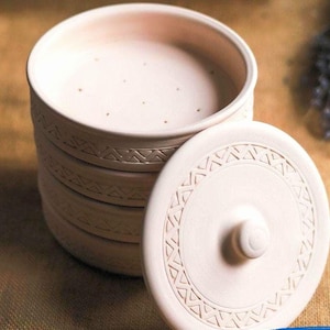 Könnte beinhalten: Ein Stapel hellbeige Keramikschalen mit passendem Deckel. Die Schalen haben ein einfaches, geometrisches Muster am Rand. Der Deckel hat einen kleinen Knopf und ein ähnliches Design. Die Schalen sind auf einer strukturierten, neutralfarbenen Oberfläche gestapelt.