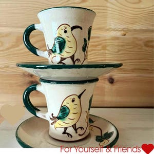 May include: Two ceramic mugs with matching saucers. The mugs and saucers are off-white with a green trim. Each mug features a hand-painted yellow bird with green accents. The text "For Yourself & Friends" is at the bottom.