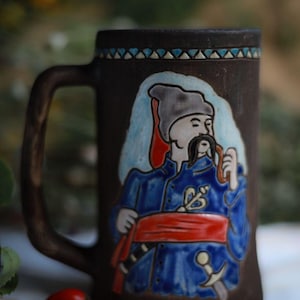 May include: A brown ceramic mug with a handle, featuring a painted image of a man in traditional Ukrainian attire. The man wears a blue coat, red sash, and a hat. The mug has a decorative border and a sword.