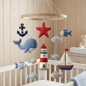 May include: A baby mobile with a nautical theme, featuring a wooden ring and hanging felt ornaments. The mobile includes a blue whale, red starfish, lighthouse, sailboat, anchor, and fish in blue and beige. It hangs above a white cot.