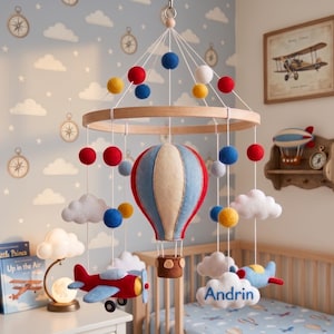 Puede incluir: Un móvil colgante para habitación infantil con un globo aerostático central de fieltro en rojo, azul y crema, rodeado de nubes de fieltro, pequeñas esferas de colores y dos aviones pequeños. Una nube blanca tiene bordado el nombre Andrin en azul.