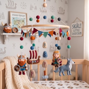 May include: A nursery mobile featuring felt figures of a Viking, a shield-maiden, a longship, a wolf, and a hammer, suspended from a wooden ring. The mobile includes colourful felt balls, triangular bunting, and felt stones with runic symbols.