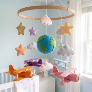 May include: A hanging mobile featuring a central felt globe, surrounded by plush felt airplanes in orange, purple, blue, and pink. The mobile includes various felt clouds in pink and grey, along with stars in gold, yellow, and purple, all suspended from a circular wooden frame.