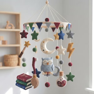 May include: A hanging mobile featuring felt shapes including a central gray owl with glasses, a crescent moon, stars, a lightning bolt, a stack of three books, a broomstick, and a potion bottle. The circular frame is decorated with colorful felt pennants.