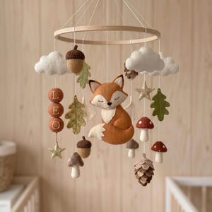 May include: A baby mobile with a woodland theme, featuring a felt fox, mushrooms, acorns, leaves, and clouds. The mobile includes the name "LEO" and is suspended from a wooden ring. The colour palette includes orange, brown, white, and green.