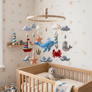 May include: A nautical-themed baby mobile featuring felt figures of a blue whale, a red crab, a grey seal, a sailboat, starfish, clouds, and waves suspended from a circular wooden frame with white string and small wooden beads.