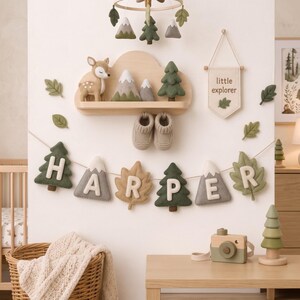 May include: A nursery wall display featuring a cloud-shaped wooden shelf holding a felt deer, mountains, and a tree. Below, a felt banner spells HARPER with tree, mountain, and leaf shapes. A little explorer wall hanging and baby booties are included.