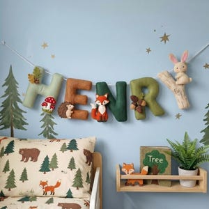 May include: A felt letter garland spelling HENRY in shades of green and brown, decorated with plush woodland animals including a hedgehog, fox, and rabbit, plus mushrooms and acorns, suspended on a string against a light blue wall.