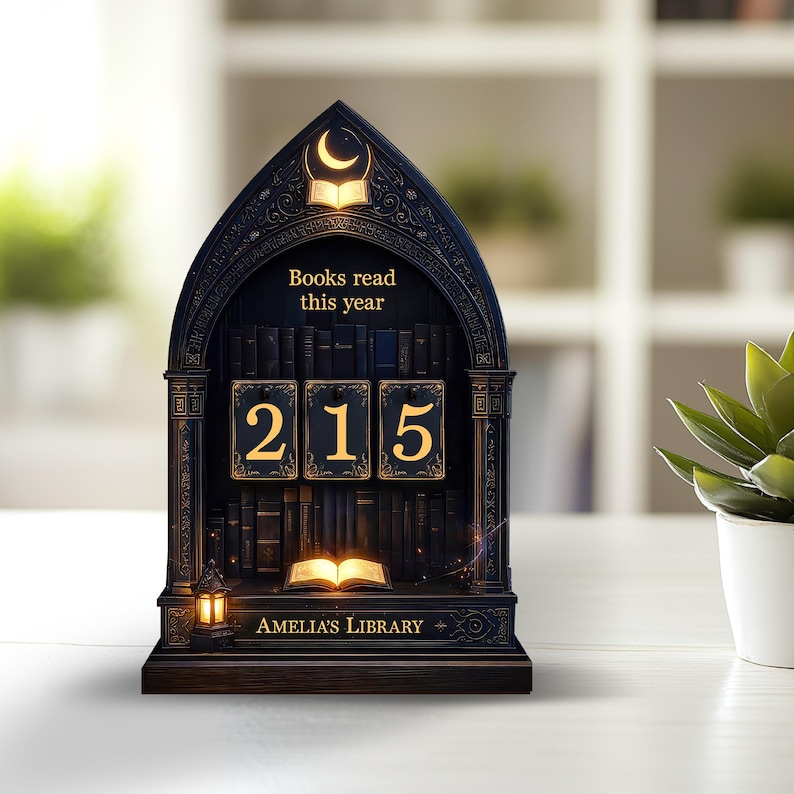 May include: A dark wooden bookshelf-shaped book counter with the text "Books read this year" and the number "215" displayed on gold-framed blocks. The base reads "Amelia's Library". A small lantern and an open book are also featured.