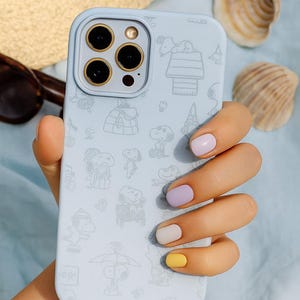 Puede incluir: Funda de teléfono azul claro con un patrón repetido de ilustraciones de Snoopy. La funda tiene un recorte para la cámara con anillos dorados. El fondo incluye un sombrero de paja, gafas de sol y conchas marinas.