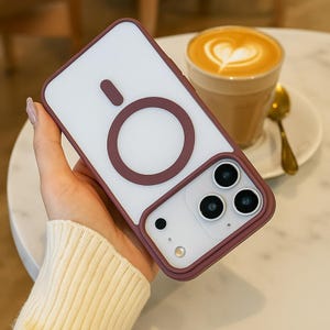 May include: A protective phone case with a white back and a burgundy-colored frame and accents. The case features a circular design on the back and a camera cutout with three lenses. The case is held in a person's hand.