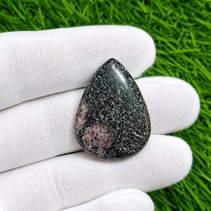 May include: A teardrop-shaped cabochon gemstone featuring a dark black base colour with intricate white speckled patterns and a distinct, rounded patch of dusty pink mineral inclusion visible on the lower left side of the polished surface.
