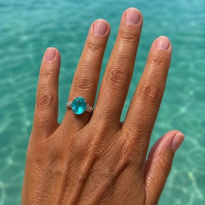 Op de afbeelding: Een zilveren ring met een grote, ovaal geslepen, helder turkooisblauwe edelsteen in het midden, geflankeerd door twee kleinere, heldere, rond geslepen accentstenen aan weerszijden, gemonteerd op een dunne, gepolijste metalen band.