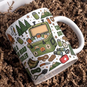 May include: A white ceramic mug featuring a colorful camping-themed pattern including a green cooler, pine trees, a campfire, hiking boots, a first aid kit, and various outdoor gear icons, with the text "CAMPING DAD" and "TAKE A BEAR ROAD COOLER".