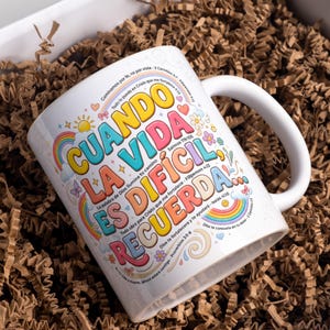 Puede incluir: Taza de cerámica blanca con asa curva, con texto colorido en español: "CUANDO LA VIDA ES DIFICIL, RECUERDA." El diseño incluye arcoíris, flores y citas inspiradoras. La taza se presenta en una caja de regalo con papel arrugado marrón.