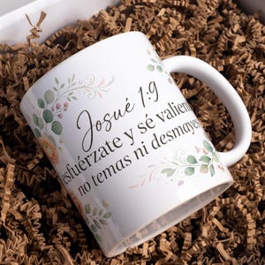May include: White ceramic mug with a curved handle, featuring floral accents in soft pink and green. The mug has text in Spanish. The mug is presented in a gift box with brown paper filler.