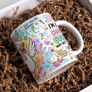 Puede incluir: Una taza de cerámica blanca con un estampado de dibujos animados de gansos coloridos y textos que incluyen "JUST CHILLING. SERIOUSLY.", "I'M A CHAOS GOOSE WHY NOT?" y "SQUAD GOALS: CARBS & CHAOS.", junto a pequeñas ilustraciones de maracas, flores y caras sonrientes.