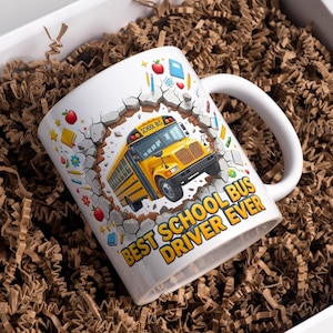 Può includere: Una tazza in ceramica bianca con la grafica di uno scuolabus giallo che emerge da un design a muro incrinato. La tazza include icone scolastiche colorate come mele, matite e libri, con il testo BEST SCHOOL BUS DRIVER EVER stampato in giallo.