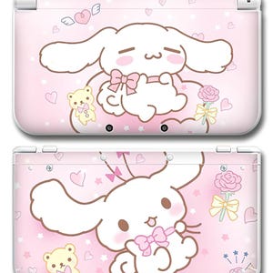 May include: A pink Nintendo 3DS case with a cartoon character design. The case features a white rabbit-like character with a pink bow, hearts, stars, and a teddy bear. The edges of the 3DS case are white.