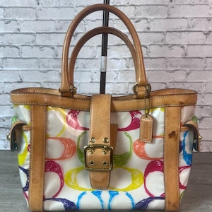 May include: A white handbag featuring a colorful multi-hued circular pattern, tan leather trim, two top handles, and a central vertical leather strap with a gold-tone metal buckle closure. A small rectangular leather tag hangs from one handle.
