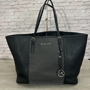 May include: Black tote bag with silver-tone stud accents and handles. The bag features the text "MICHAEL KORS" and a silver "MK" logo charm. The bag has a rectangular shape and is made of a smooth material.