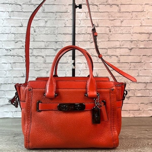 May include: A bright orange leather handbag featuring dual top handles, a detachable shoulder strap, and a dark metal turn-lock closure on the front. The bag includes a matching leather luggage tag and dark metal hardware accents throughout.