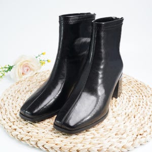 以下が含まれることがあります： スクエアトゥとブロックヒールが特徴の黒いアンクルブーツのペア。ブーツは光沢のある仕上がりで、背面にジッパーが付いています。白い花と緑のある、織られた円形のマットの上に置かれています。