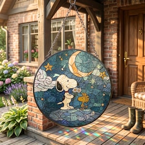 Puede incluir: Panel de vidriera redondo con Snoopy y Woodstock bajo una luna creciente y estrellas. La obra de arte utiliza tonos azules, dorados y blancos, con un borde negro y una cadena para colgar. El panel se exhibe en un porche.