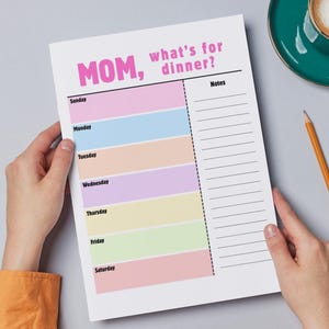 May include: A weekly meal planner with the text "MOM, what's for dinner?" is held by hands. The planner has colorful sections for each day. A cup of coffee and pencils are nearby.