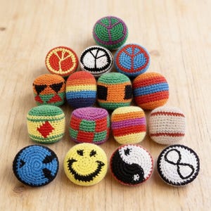 May include: A collection of colorful, handmade crochet balls. The balls feature various designs, including a smiley face, peace sign, yin-yang symbol, and geometric patterns. The balls are arranged on a light wooden surface.