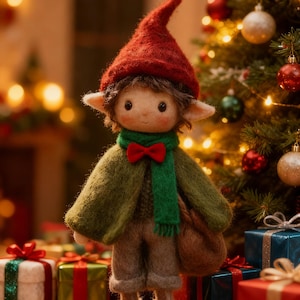 Op de afbeelding: Een handgemaakte elfenpop met een rode puntmuts, groene sjaal en olijfgroene jas staat voor een kerstboom. De pop heeft een bruine tas en is omringd door ingepakte cadeaus. De scène roept een feestelijke sfeer op.