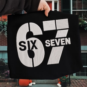 Könnte beinhalten: Schwarze Tragetasche mit dem gestickten Text "SIX SEVEN" in Weiß. Die Zahl 6 ist mit dem Wort "SIX" im Inneren stilisiert. Die Zahl 7 ist mit dem Wort "SEVEN" daneben stilisiert.