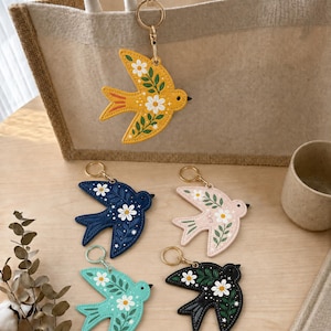 May include: Five bird-shaped keychains featuring floral patterns with white daisies and green leaves. The birds come in yellow, navy blue, light pink, mint green, and black, each attached to a gold-toned metal ring for secure attachment.