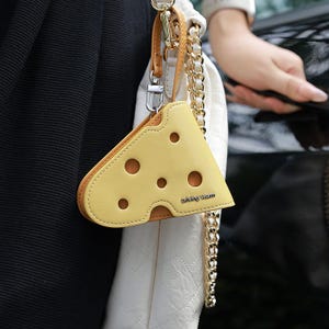 Puede incluir: Un estuche triangular de color amarillo claro con forma de cuña de queso suizo con recortes circulares, que presenta el texto "Driving Warm" en pequeñas letras plateadas. El estuche está sujeto a un clip de metal dorado y una correa de cuero amarilla.