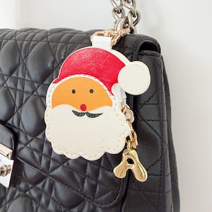 Puede incluir: Un colgante para bolso con forma de Papá Noel que presenta un gorro rojo con pompón blanco, cara naranja, nariz roja y bigote negro. El accesorio está sujeto a un bolso negro acolchado con cadena plateada y tiene un dije dorado con la letra A.