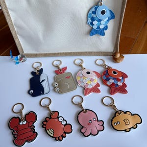 Op de afbeelding: Een verzameling sleutelhangers met zeedieren, waaronder vissen, een kreeft, een krab en een octopus. De sleutelhangers zijn gemaakt van leer en hebben goudkleurige sleutelringen. De vissen hebben blauwe, rode en roze schubben.