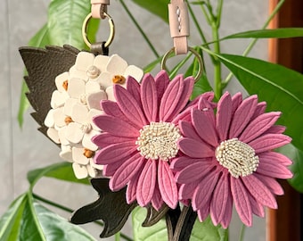 Puede incluir: Dos llaveros de cuero con forma de flor, que incluyen margaritas rosas con centros crema y un racimo blanco estilo hortensia con detalles marrones. Cada flor está unida a una anilla de metal dorado y a una correa de cuero beige claro con costuras.