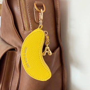 May include: A bright yellow, crescent-shaped coin purse with a textured surface and the embossed text Have a nice day. It features a gold-toned metal zip, a small gold-toned letter A charm, and a gold-toned lobster clasp attached to a brown bag.