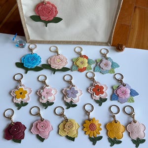 May include: A collection of seventeen floral keychains featuring various layered leather-like flower designs in colors including pink, blue, yellow, orange, purple, and red, each attached to a gold-tone metal split ring and a green leaf accent.