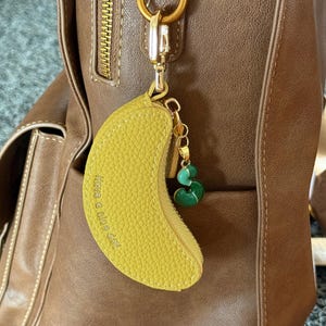 May include: A yellow banana-shaped keychain with the text "Have a nice day" is attached to a brown leather bag. The keychain has a gold-colored zipper and a green charm with the letter "S".