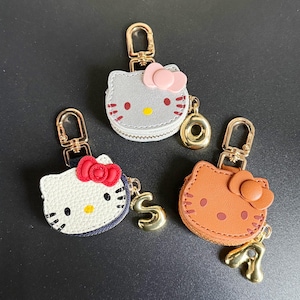 May include: Three Hello Kitty-shaped coin purses with gold-tone keychains. One is white with a red bow, another is gray with a pink bow, and the third is brown with a brown bow. Each has a gold-tone charm.
