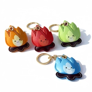 May include: A collection of four colorful keychains shaped like cartoon flames. The keychains are in orange, red, green, and blue, each with a smiling face. They have gold-colored key rings and dark brown bases.