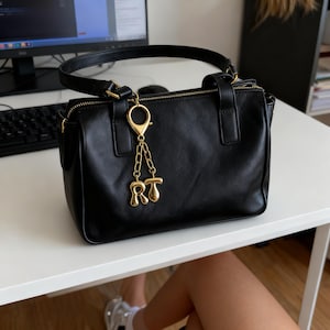 Puede incluir: Bolso de mano de cuero negro con asa superior y herrajes dorados. El bolso presenta un charm dorado con las letras "RT" colgando del asa. El bolso está sobre una superficie blanca.