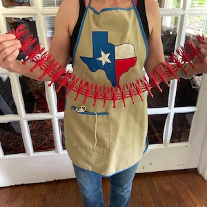May include: A person wearing a tan apron with a Texas flag design is holding a red paper chain of crawfish.