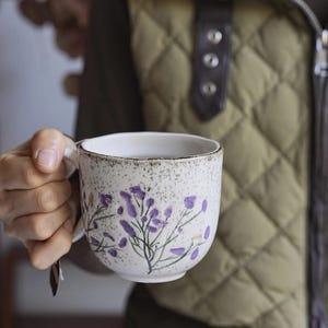 May include: A white ceramic mug with a speckled finish and a handle. The mug is decorated with purple floral sprigs. The background shows a quilted olive green vest.