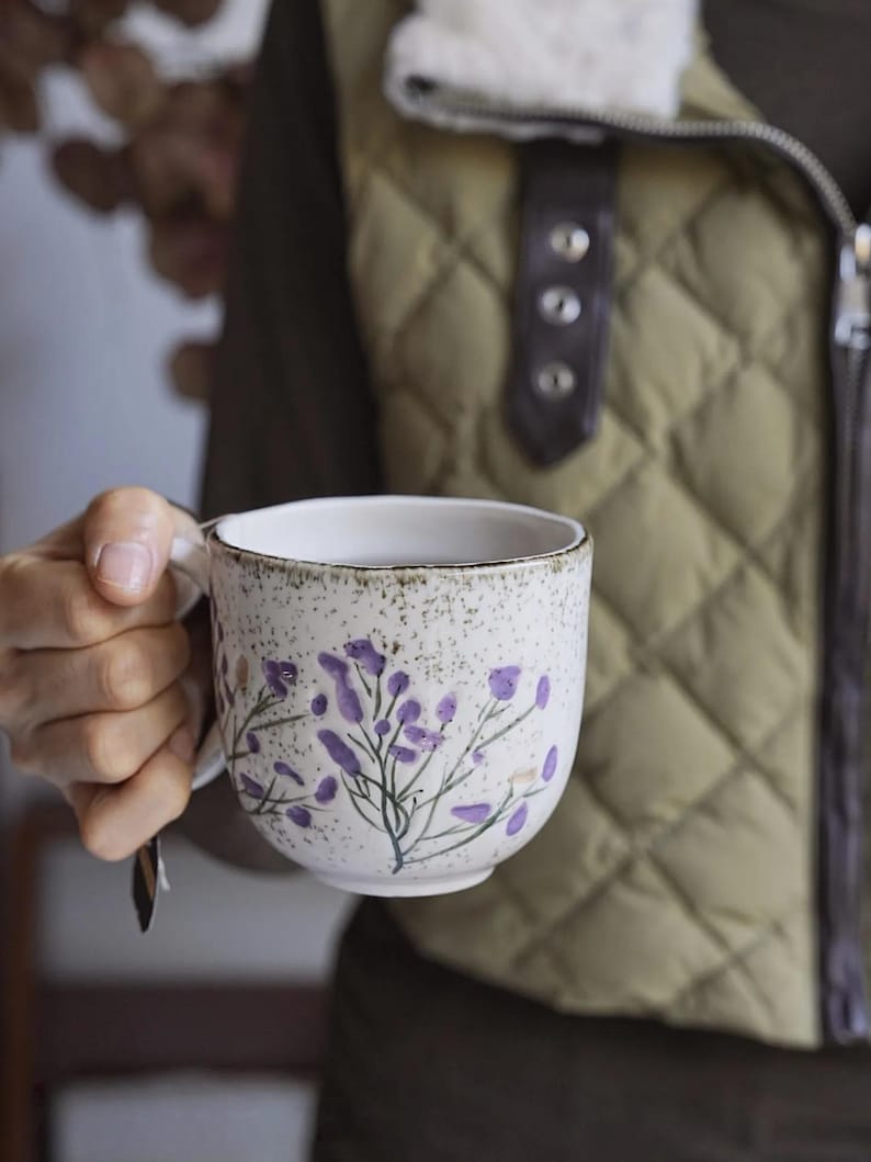 May include: A white ceramic mug with a speckled finish and a handle. The mug is decorated with purple floral sprigs. The background shows a quilted olive green vest.