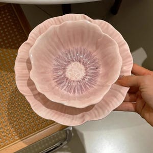 May include: A pair of pink ceramic bowls, one inside the other, designed to resemble a flower. The bowls have a textured finish and a darker pink centre. The bowls are held by a person's hand.