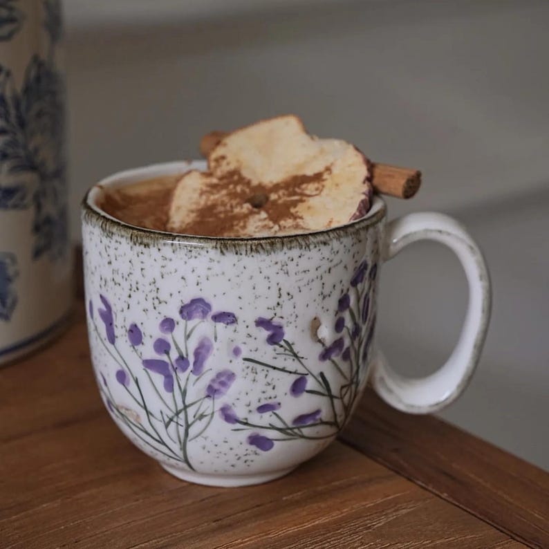 May include: A ceramic mug with a floral design, filled with a warm beverage and topped with apple slices and a cinnamon stick. The mug has a white base with purple floral accents and a speckled texture. The handle is a curved, off-white color.
